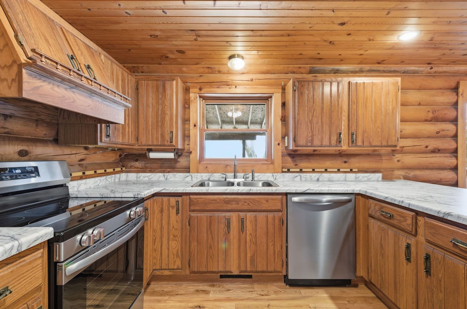 Rustic log cabin interior with wooden walls, ceiling beams, and modern stainless steel kitchen appliances.
