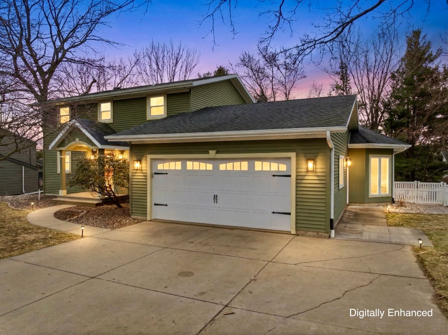 Two-story green home with white garage door, concrete driveway, and lush trees at dusk.