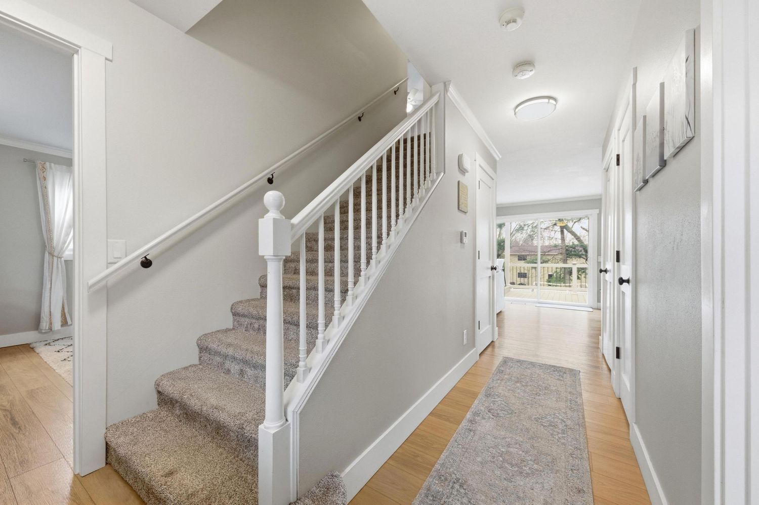 Bright hallway with white staircase, hardwood floors, gray walls, and a view to a sunlit entryway door.