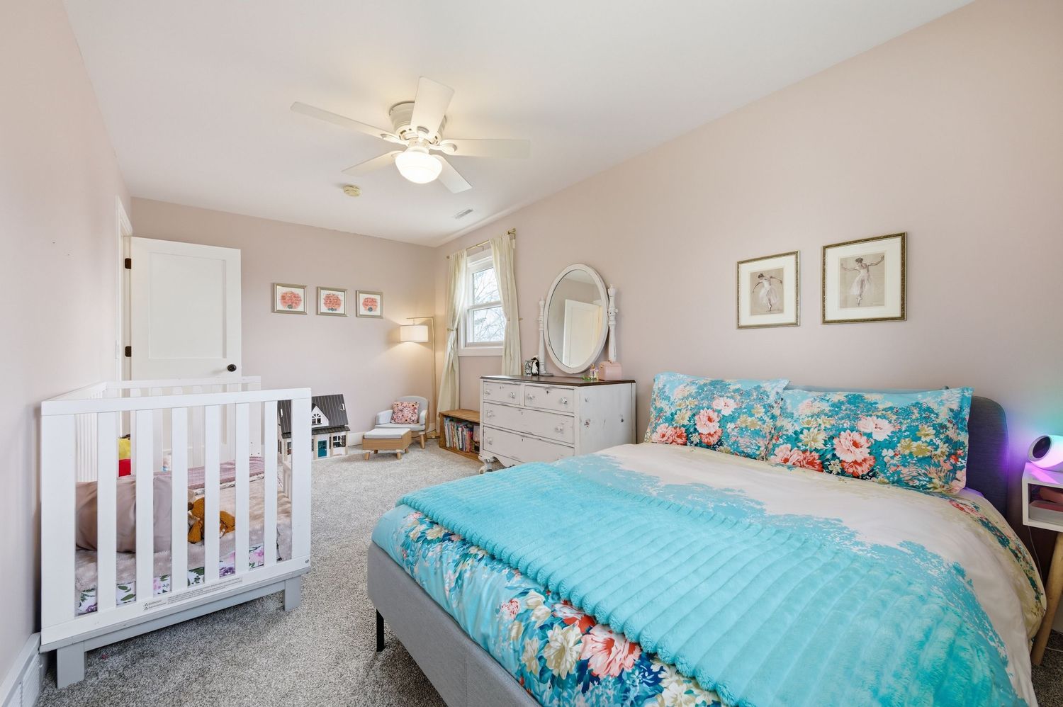 Bright bedroom with floral bedding, white crib, dresser with mirror, and ceiling fan on beige carpet.