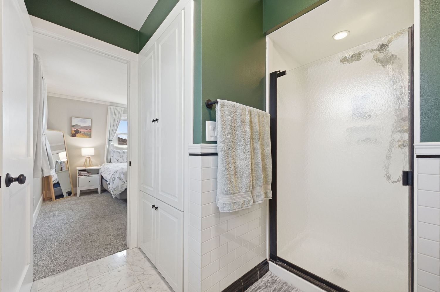 Modern bathroom with green walls, walk-in shower, white cabinetry, and an open door leading to a carpeted bedroom.