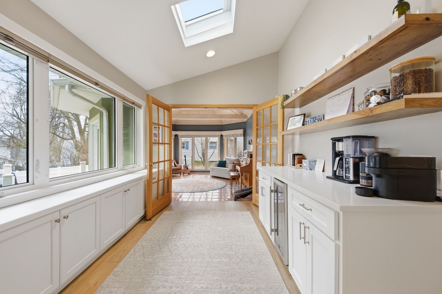 Bright sunroom with skylights, white cabinets, open wood shelving, French doors, and hardwood floors leading to a cozy living area.