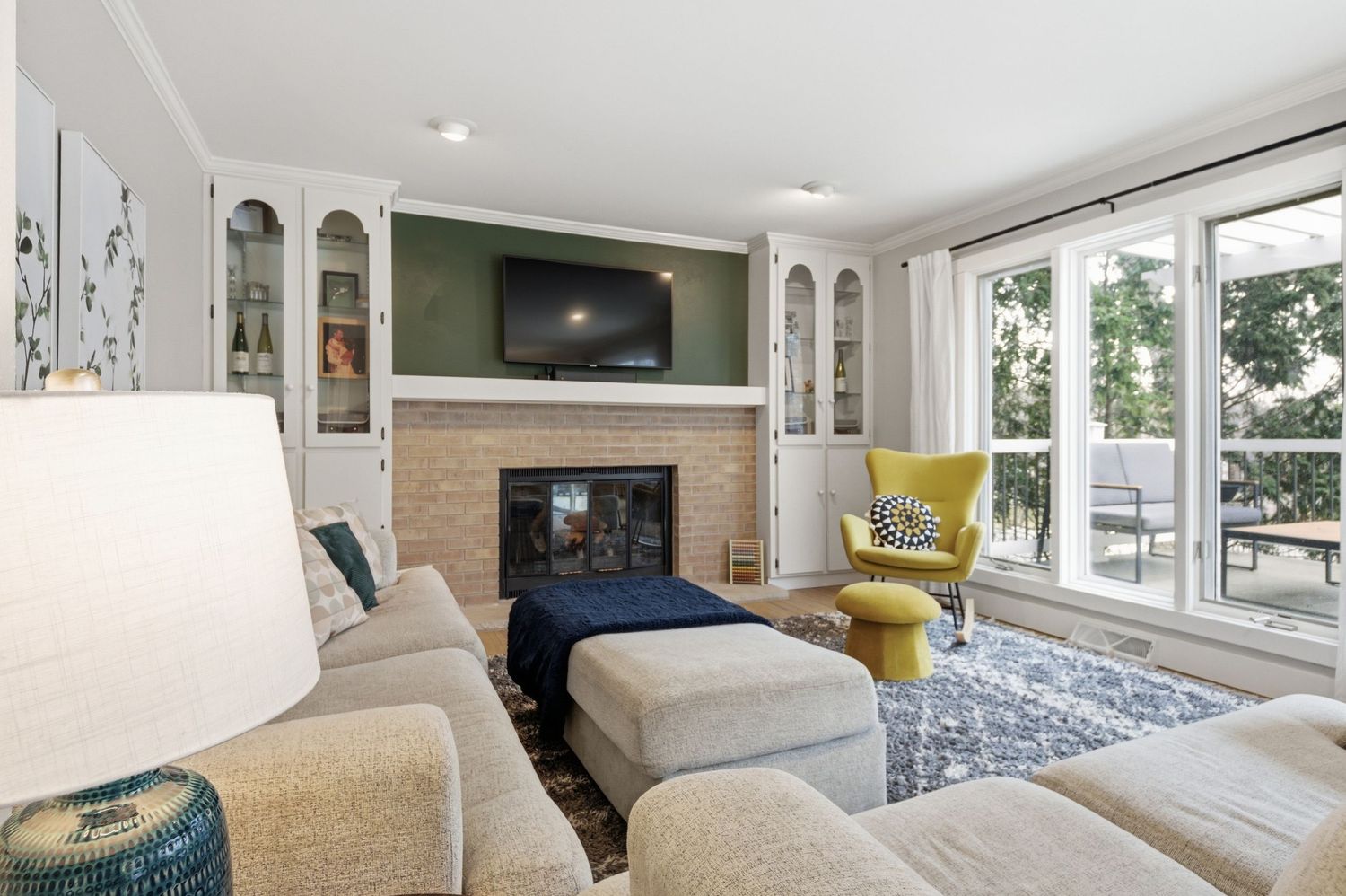 Cozy living room with built-in shelves, brick fireplace, wall-mounted TV, neutral sofas, yellow accent chair, and patio access.