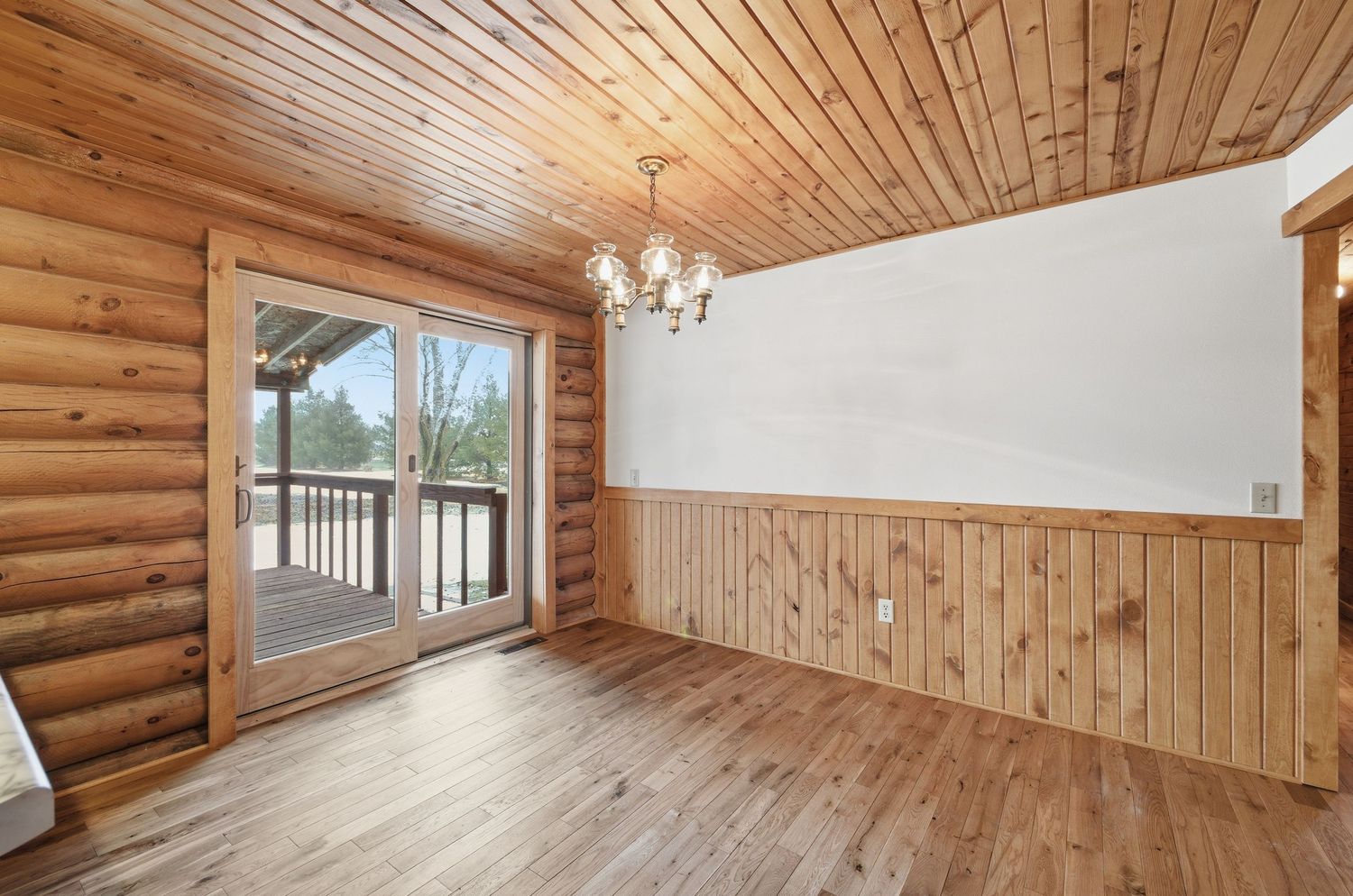 Rustic log cabin interior with wooden ceiling panels, exposed beams, and natural lighting through large windows.