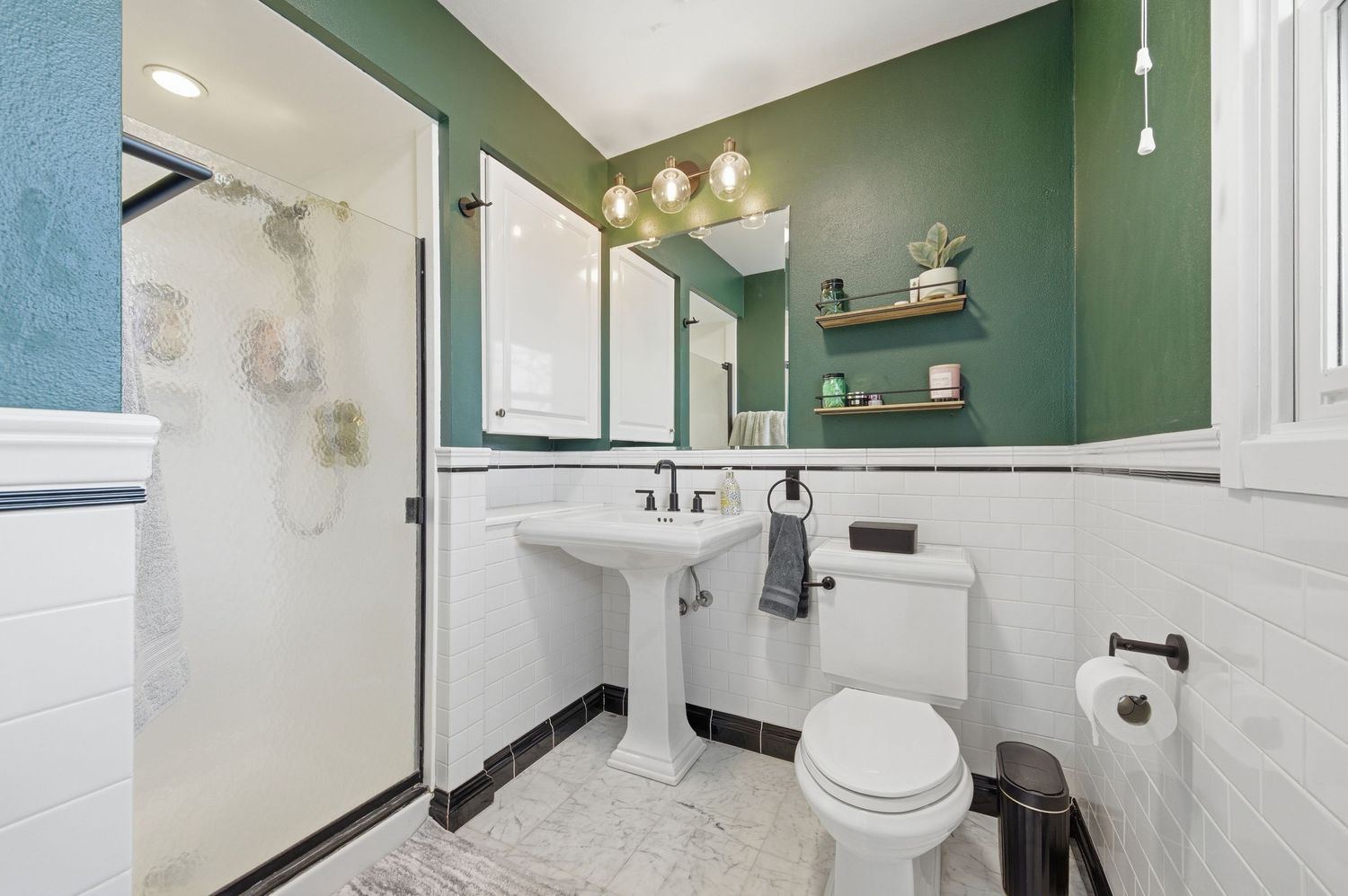 Modern bathroom with green walls, white pedestal sink, toilet, walk-in shower, and marble tile flooring.