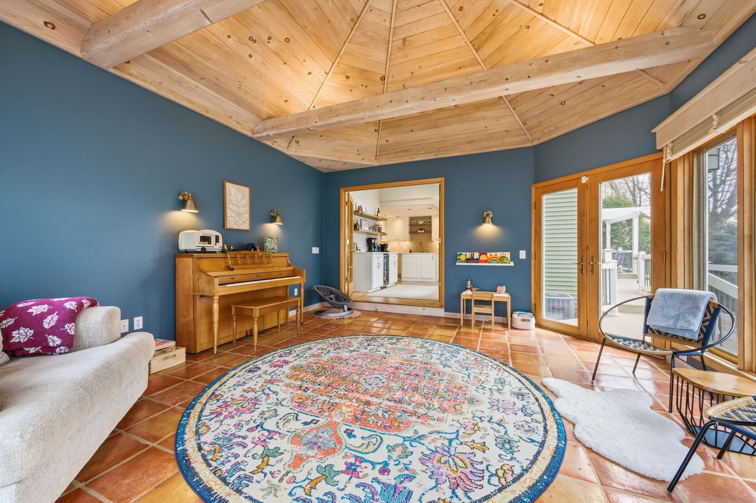 Cozy living room with blue walls, wood beam ceiling, terracotta tile floors, round floral rug, upright piano, and French doors.