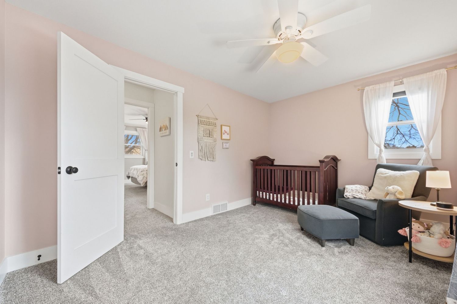 Bright nursery with pink walls, gray carpet, dark wood crib, ceiling fan, and an open door leading to an adjacent room.