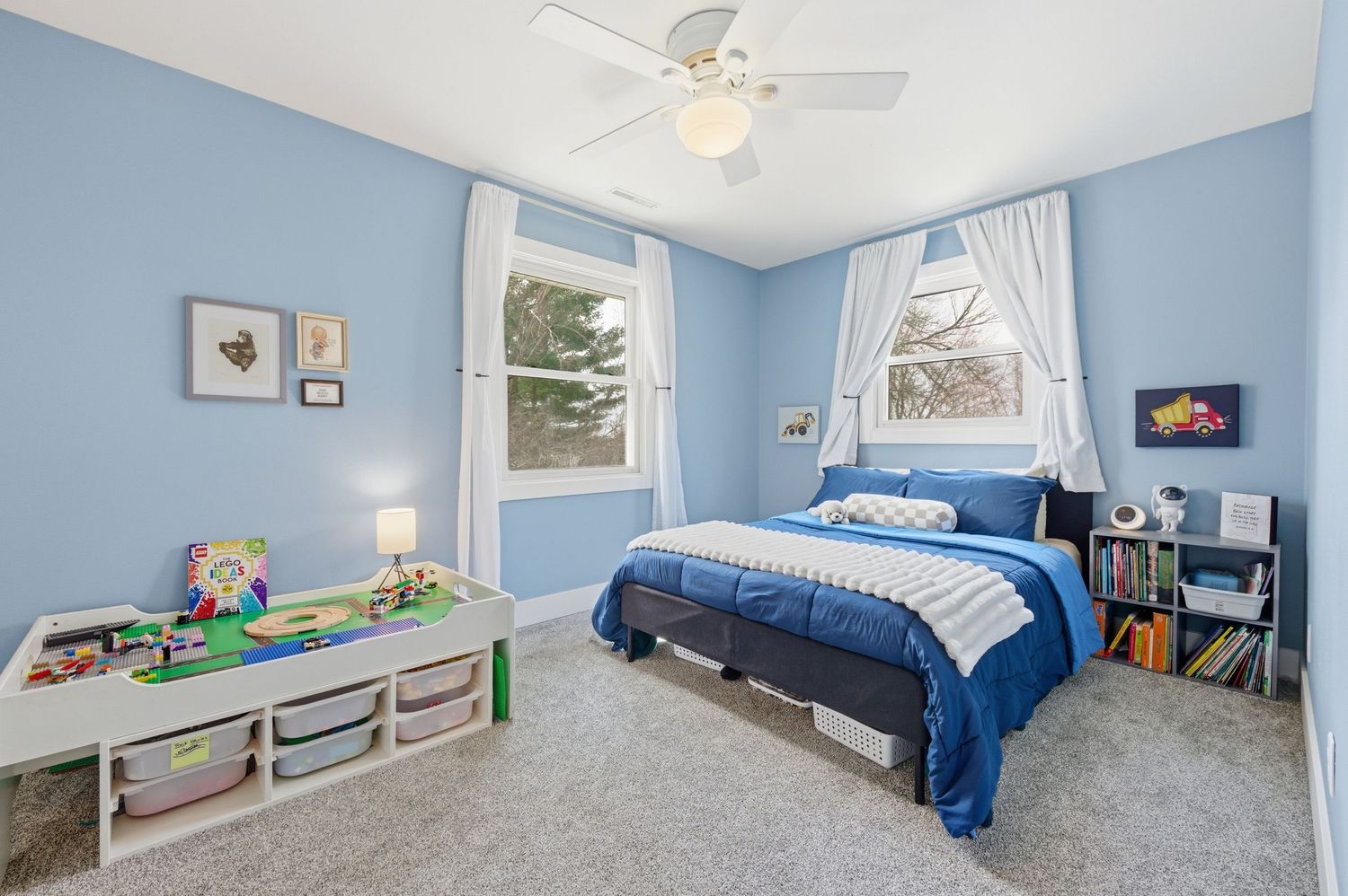Blue kids bedroom with carpet, ceiling fan, toy storage, bookshelf, and blue bedding near two bright windows.