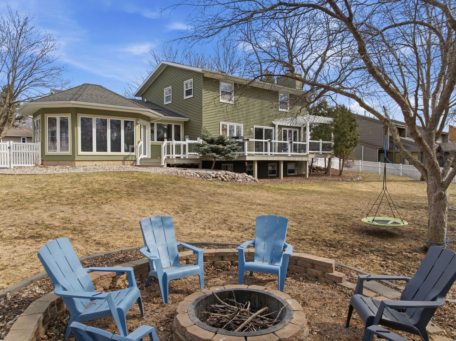 Backyard patio with blue Adirondack chairs around a stone fire pit, deck, and large green home.