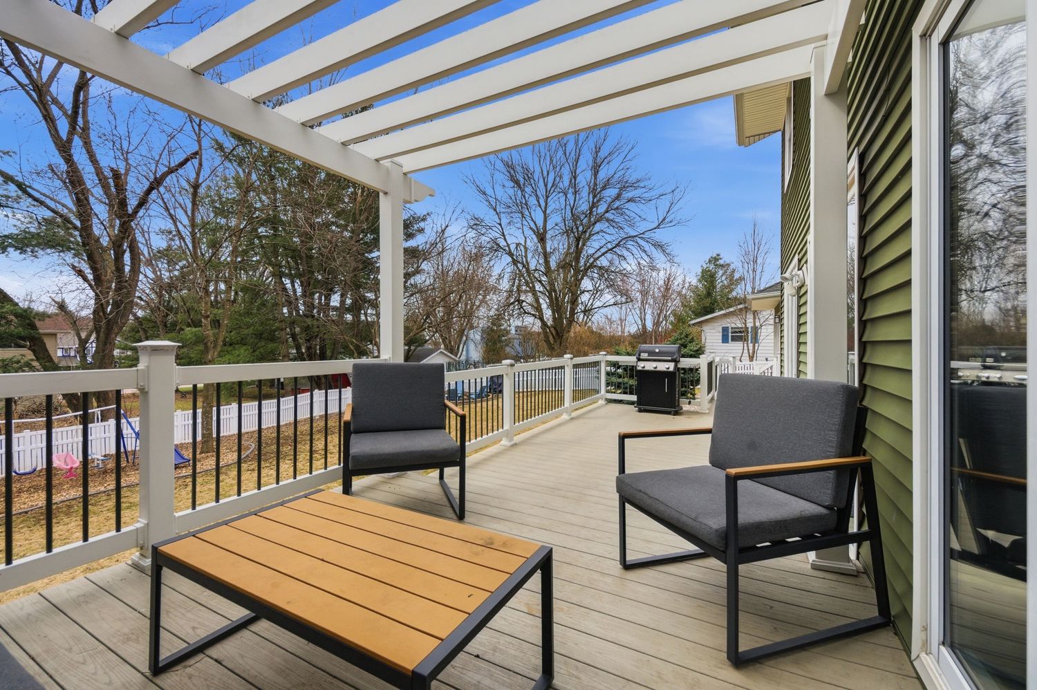 Spacious outdoor deck with modern patio furniture, pergola, and white railings overlooking a tree-lined backyard.