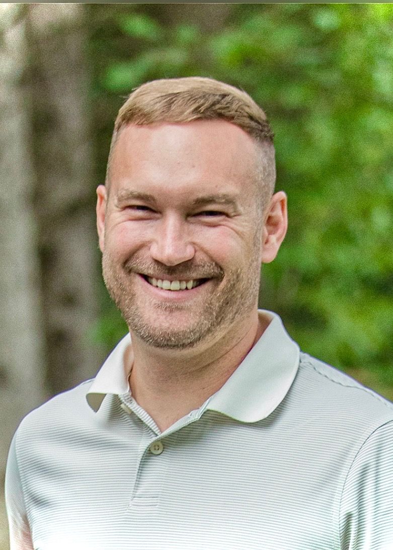 Person in white polo shirt smiling warmly outdoors with green foliage in background in Marshfield Wisconsin