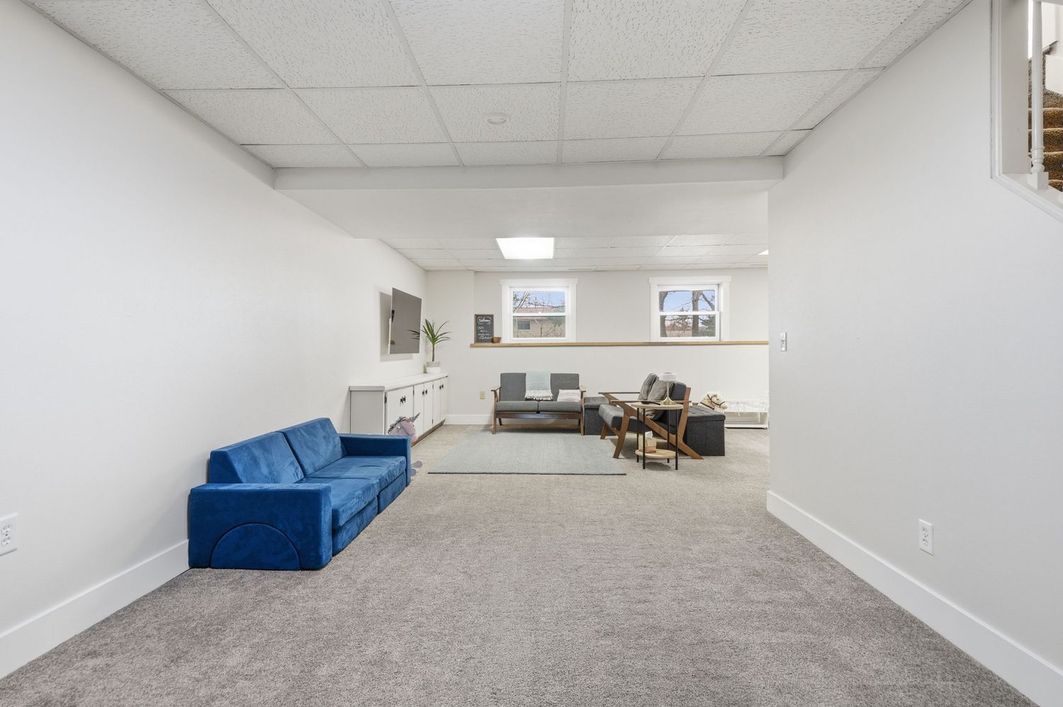 Spacious basement rec room with gray carpet, white walls, blue velvet sofa, and drop ceiling with recessed lighting.