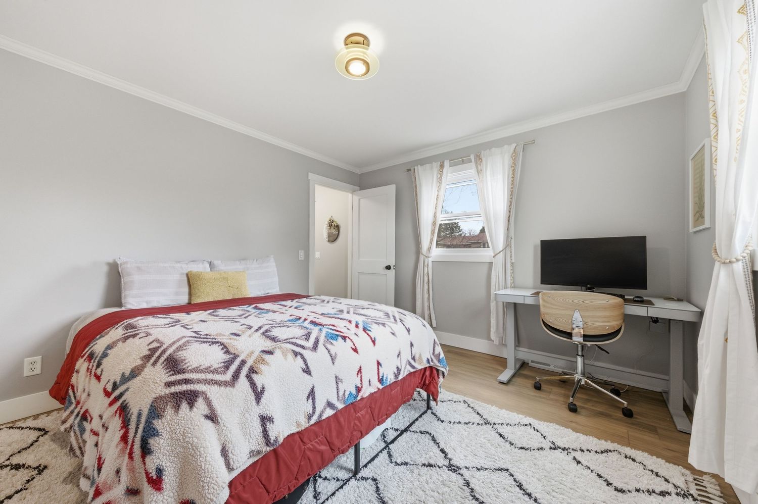 Cozy bedroom with a patterned quilt bed, white walls, crown molding, a desk with monitor, and a area rug on hardwood floors.