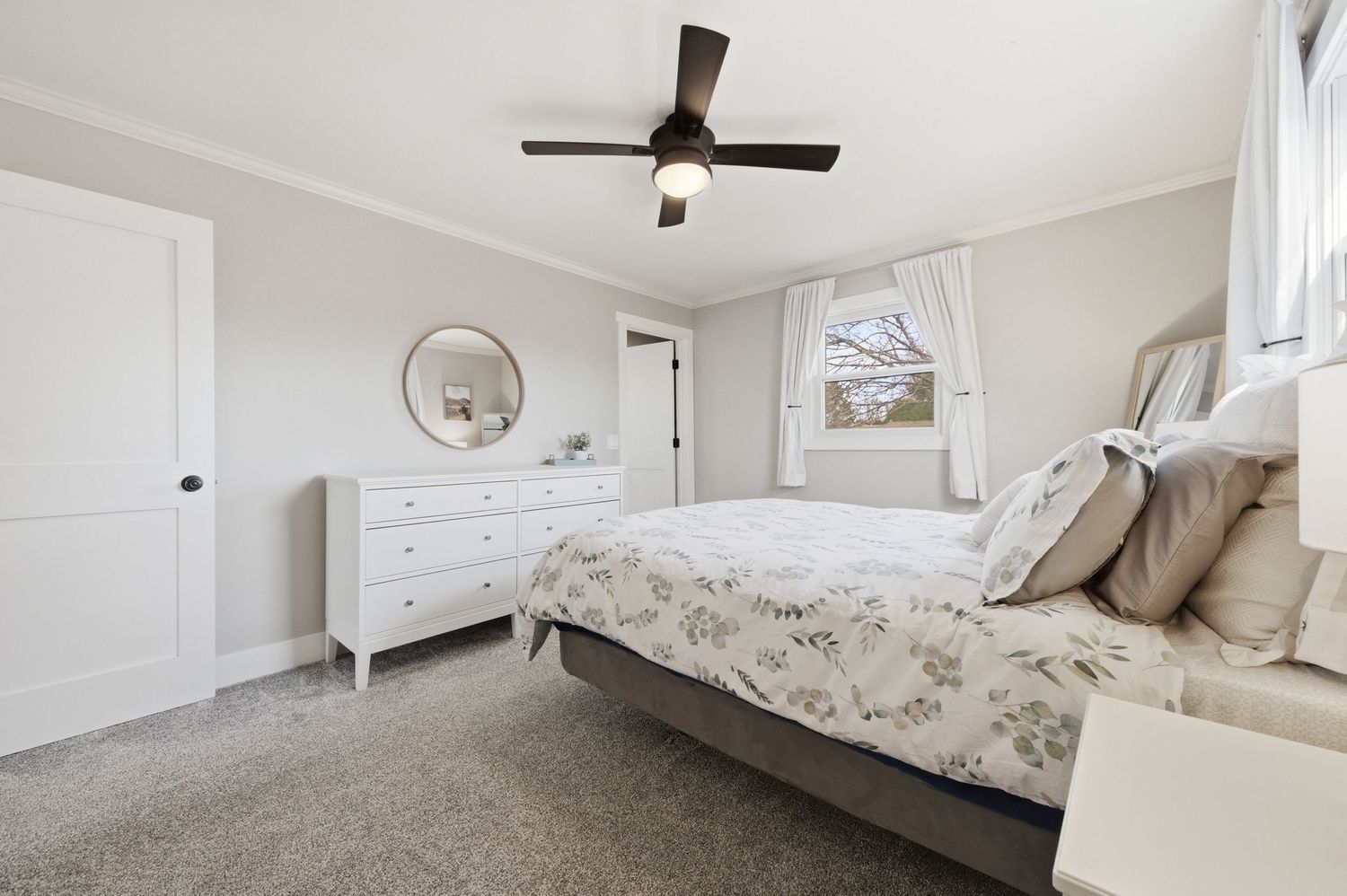 Bright bedroom with floral bedding, white dresser, round mirror, ceiling fan, and neutral carpet flooring.