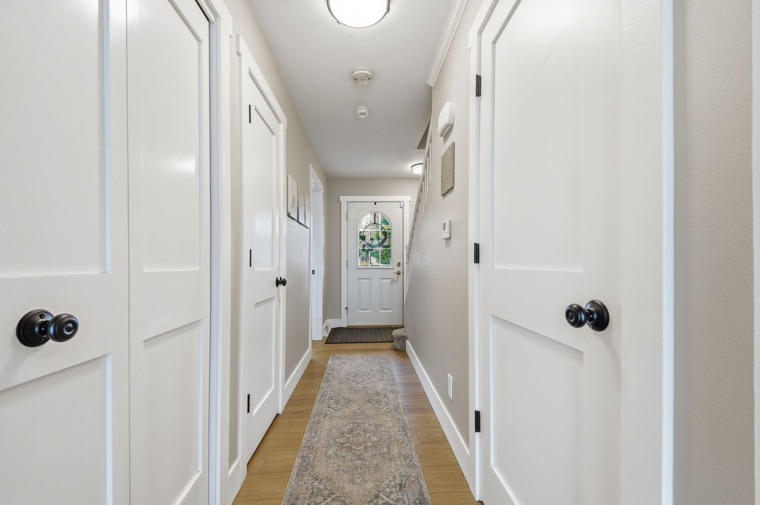 Bright narrow hallway with white paneled doors, black knobs, a patterned runner rug, and a white entry door at the end.