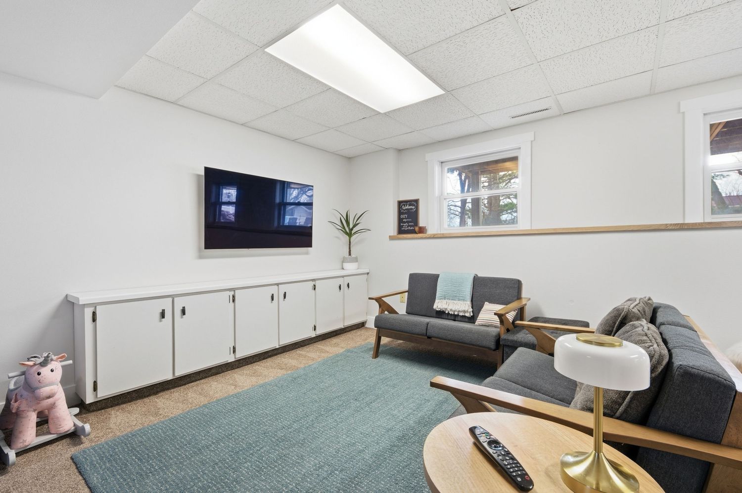 Modern basement living room with gray sofa, wall-mounted TV, white cabinets, teal rug, and natural light from window.