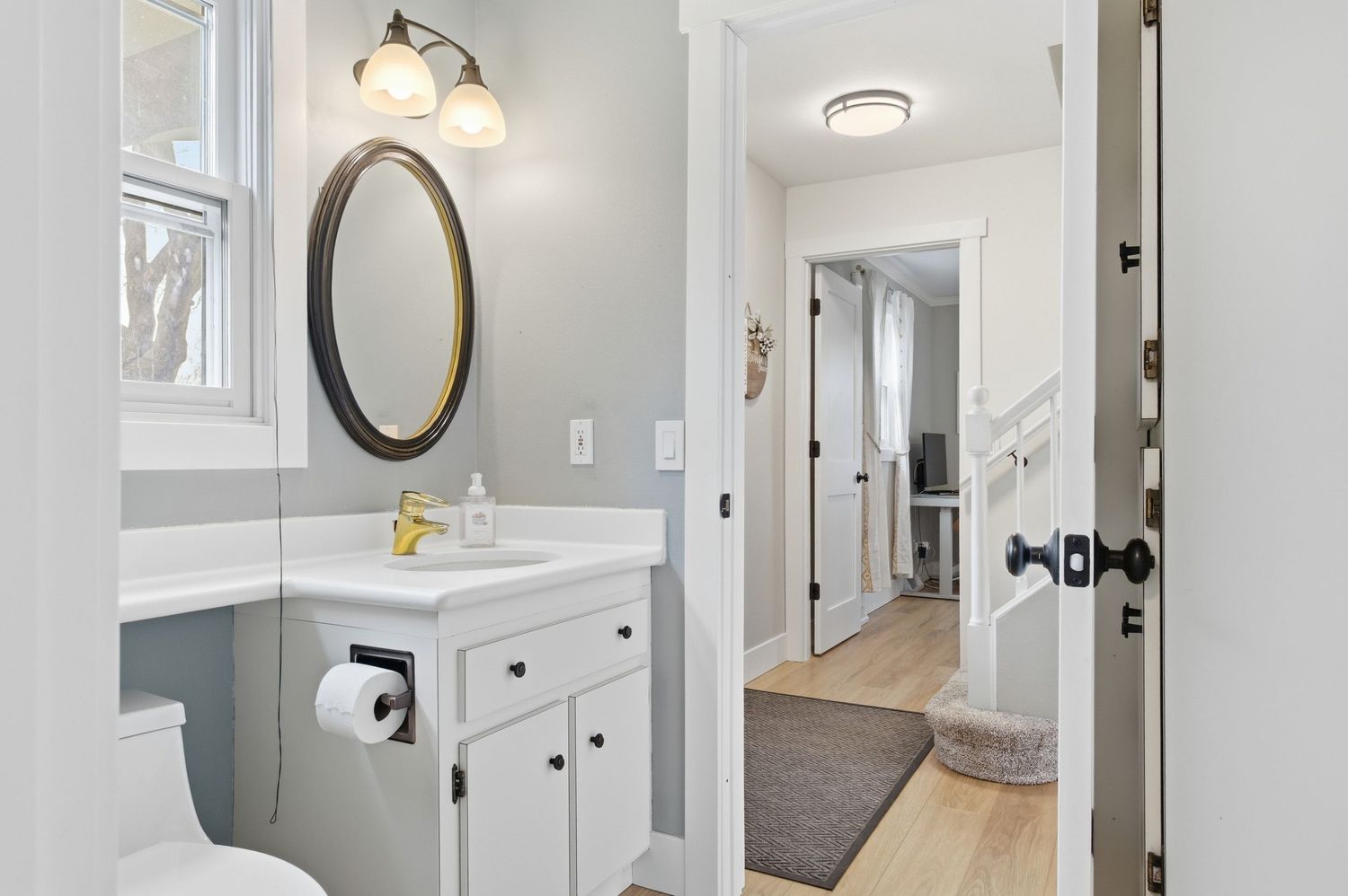 Modern bathroom with white vanity, oval mirror, and open door revealing hardwood hallway beyond.