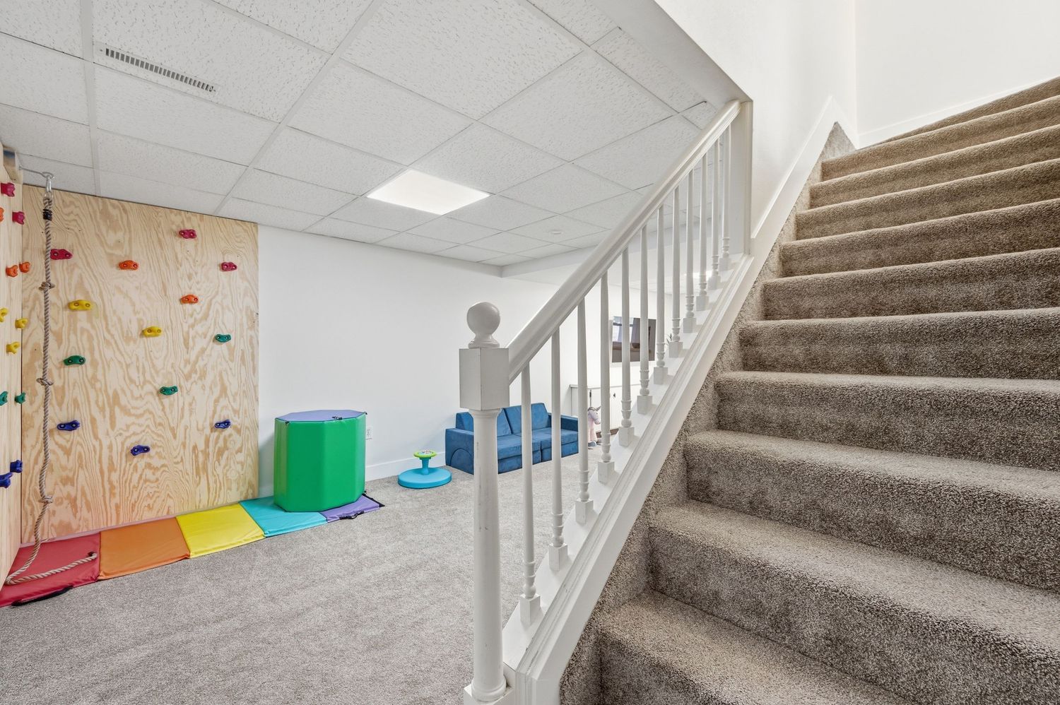 Basement playroom with carpet stairs, white railing, colorful climbing wall, and kids play area with foam furniture.