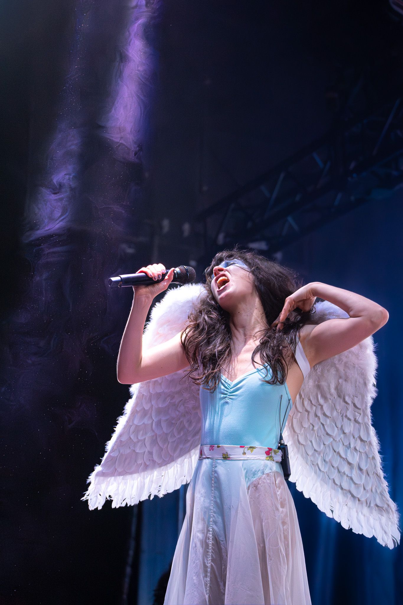 A singer in angel wings and blue dress performs passionately on stage with microphone under dramatic purple lighting.