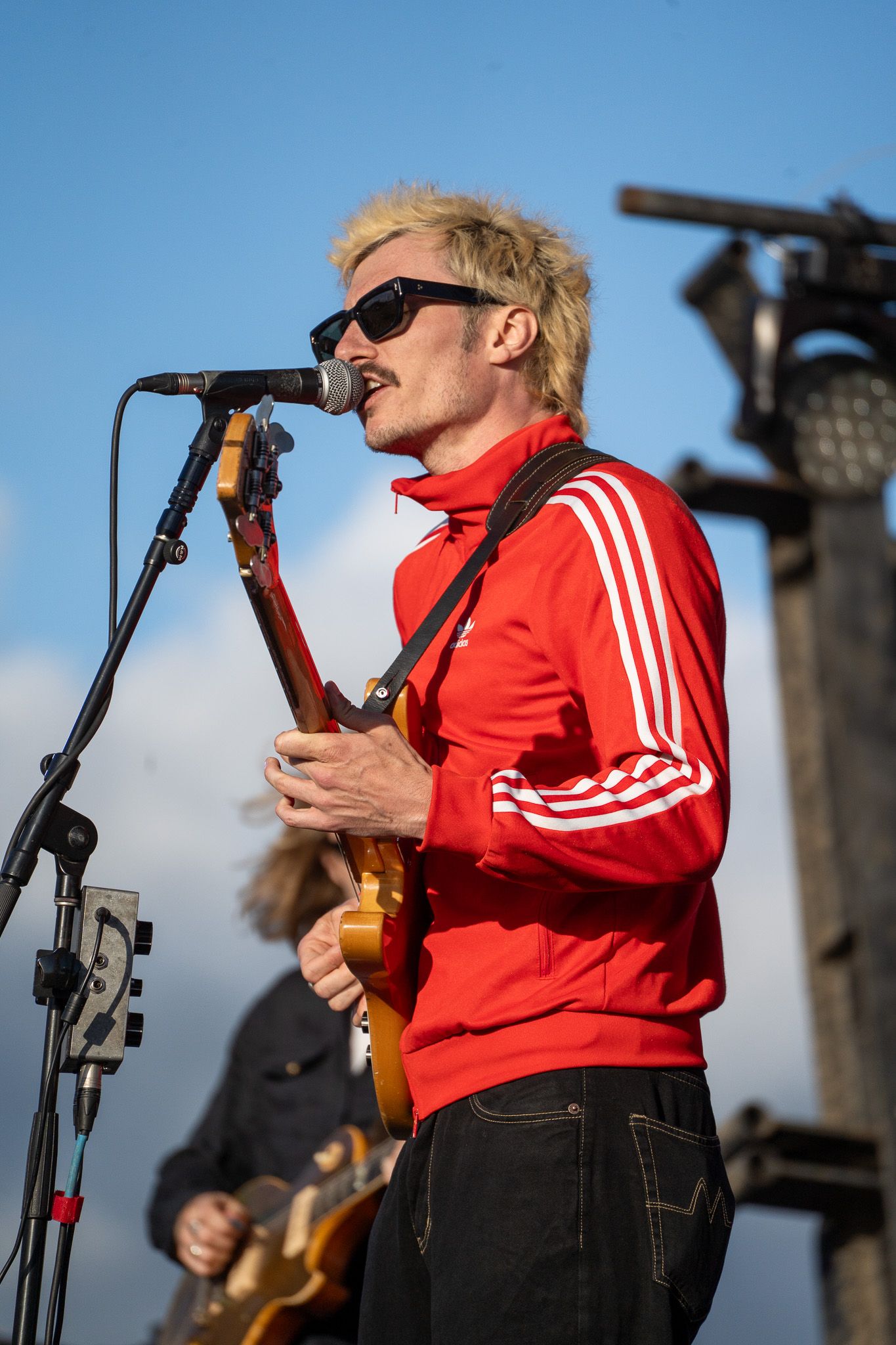 Musician in red Adidas jacket and sunglasses plays guitar and sings into microphone at outdoor concert.