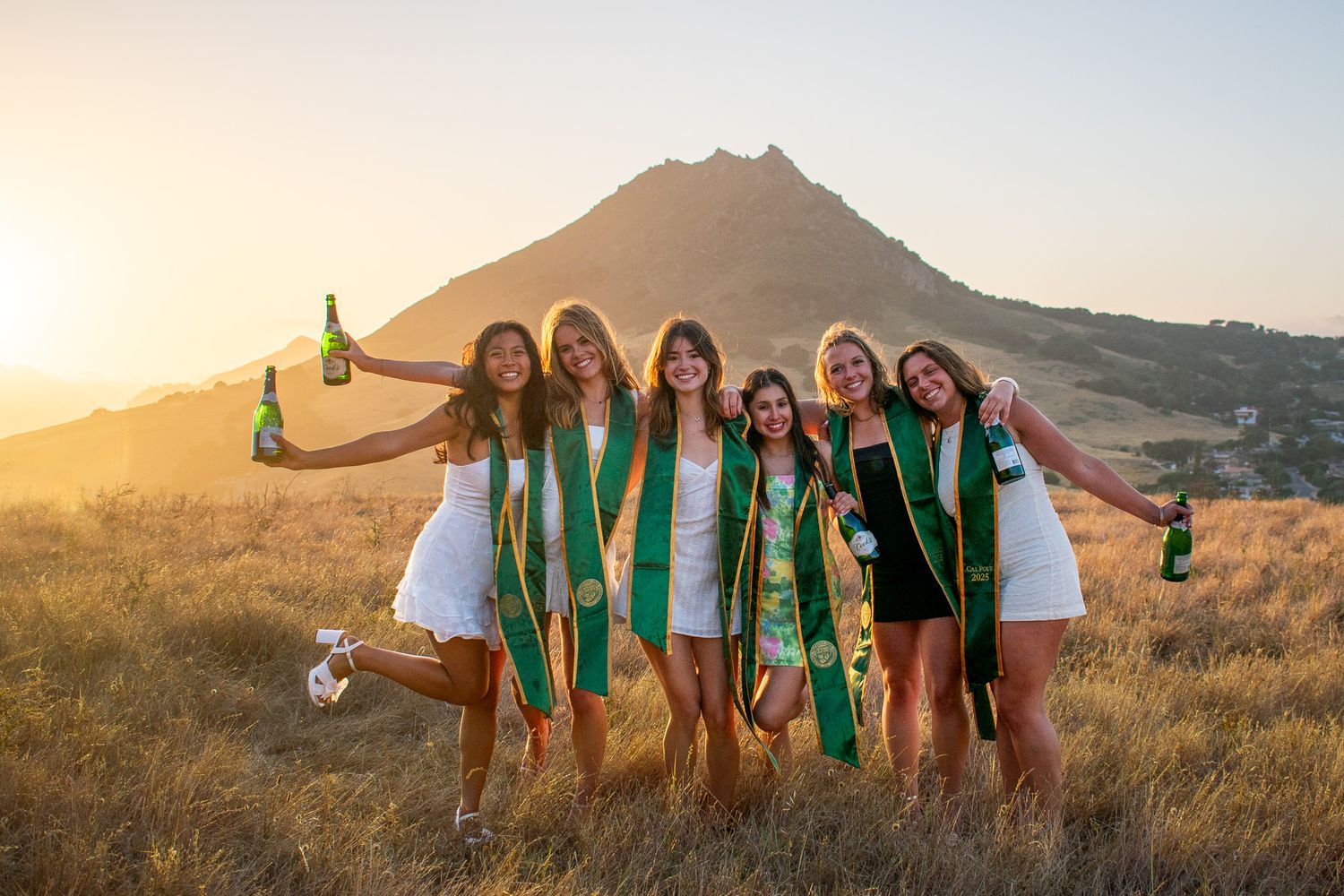 Graduates in white dresses and green stoles celebrate at sunset with mountain backdrop.