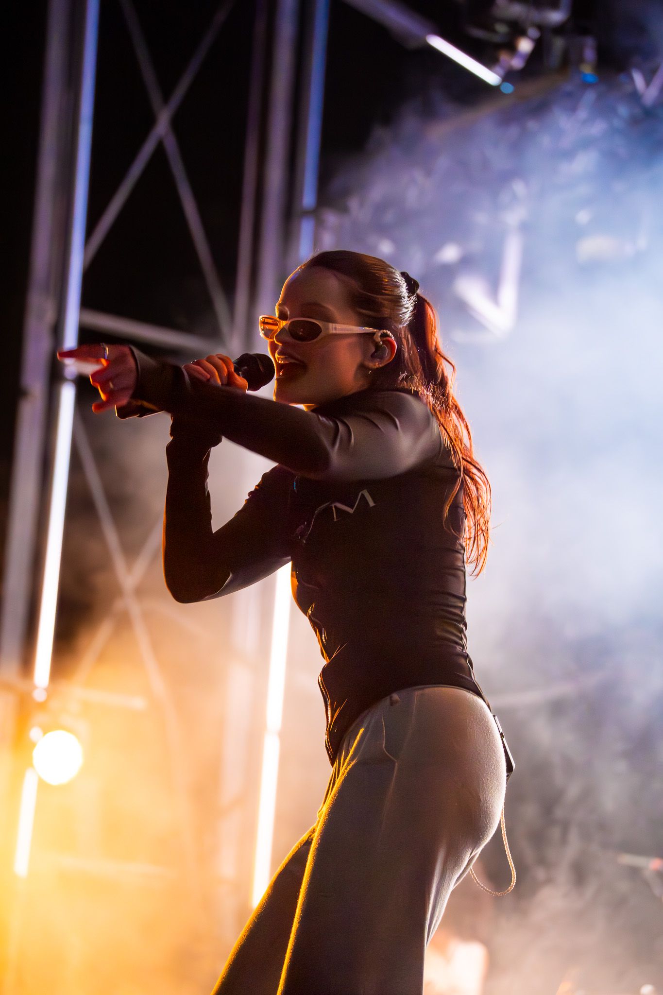 Female singer performing on stage with microphone, wearing sunglasses and black bodysuit under dramatic concert lighting.