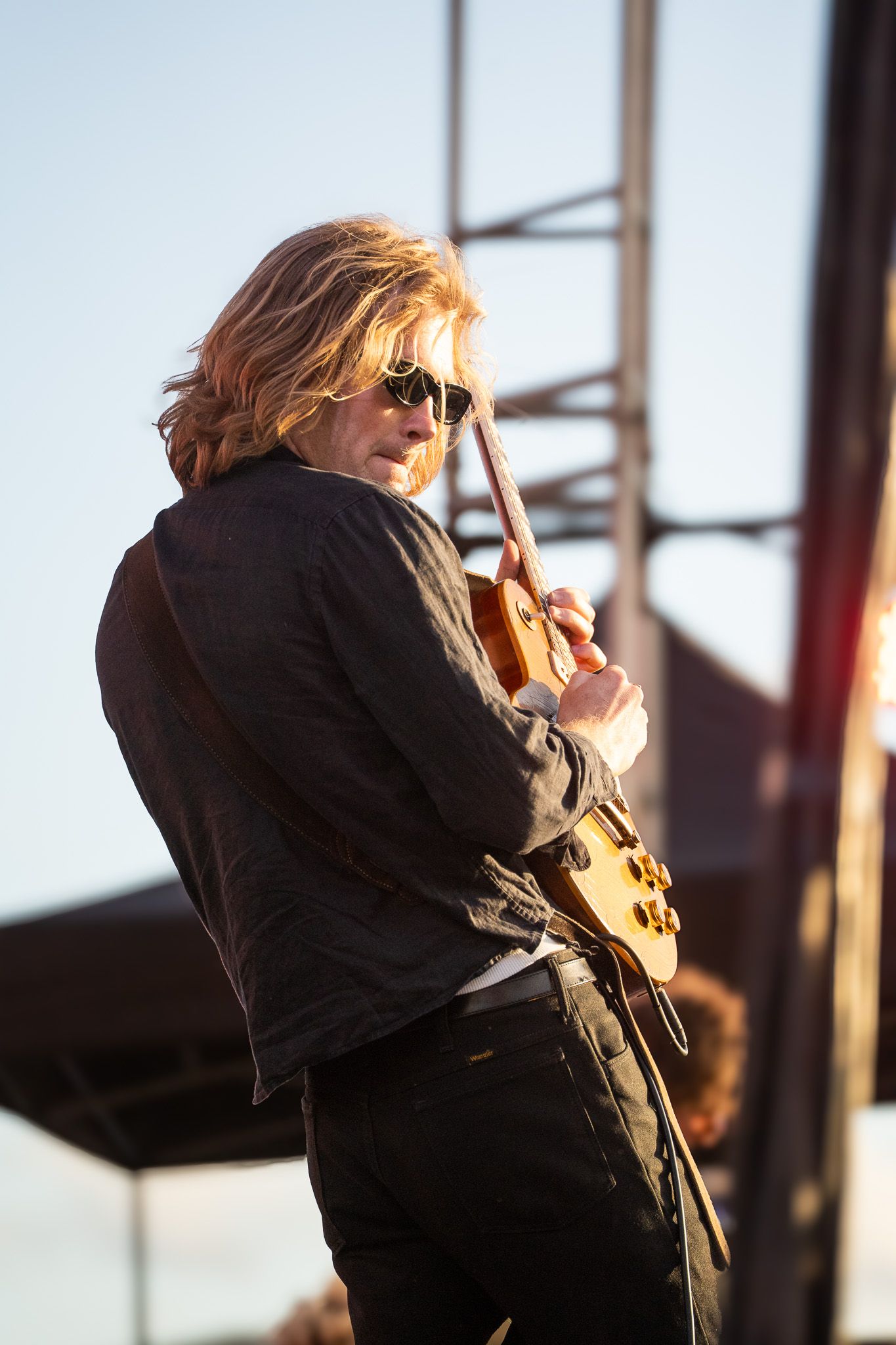 Blonde guitarist in sunglasses and black jacket plays electric guitar on outdoor stage in golden sunlight.