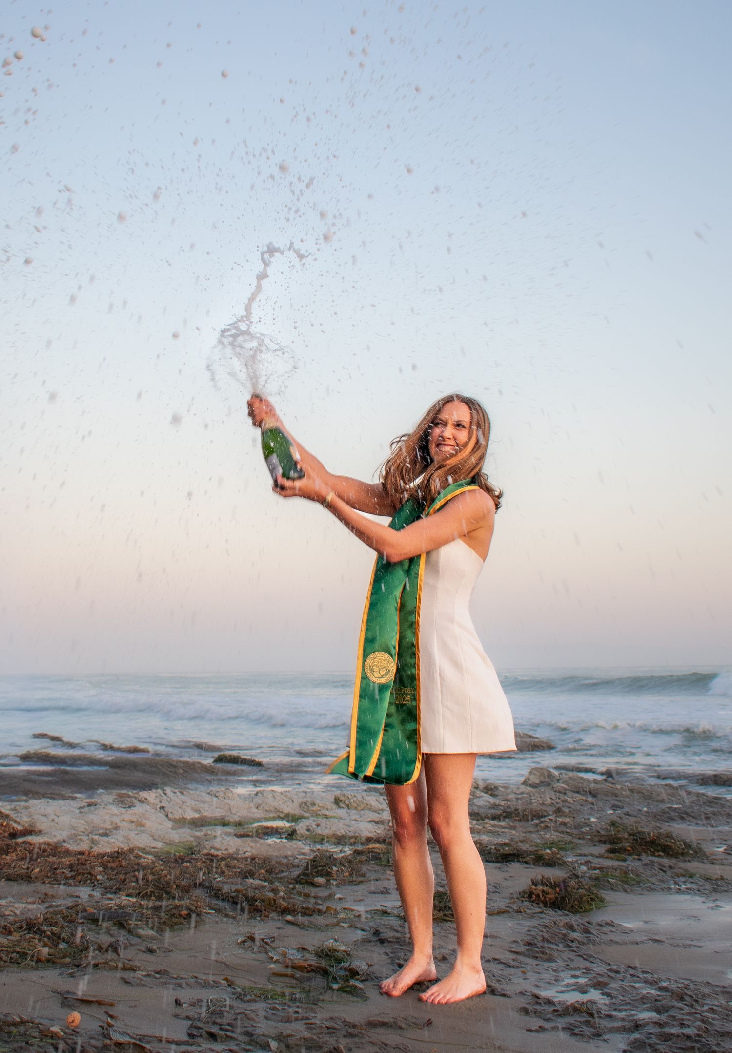 Friends celebrate on rocky beach at sunset wearing graduation attire.