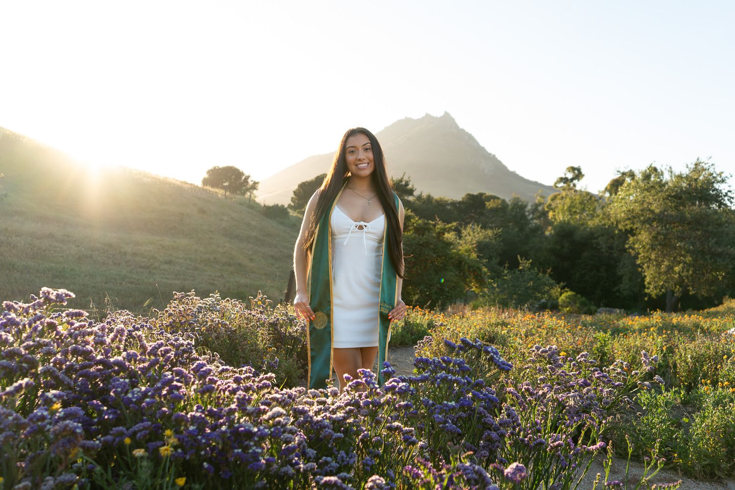 Woman in white dress and green cardigan standing among purple wildflowers with sunlit mountain backdrop.