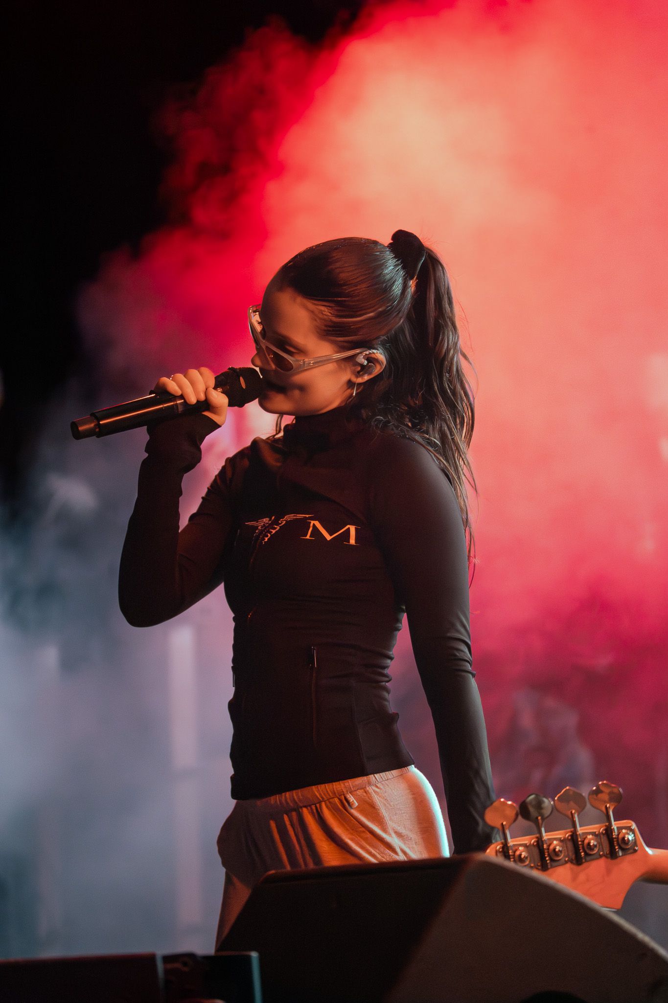 Female singer performing on stage with microphone, wearing black top and silver shorts, with red smoke lighting behind her.