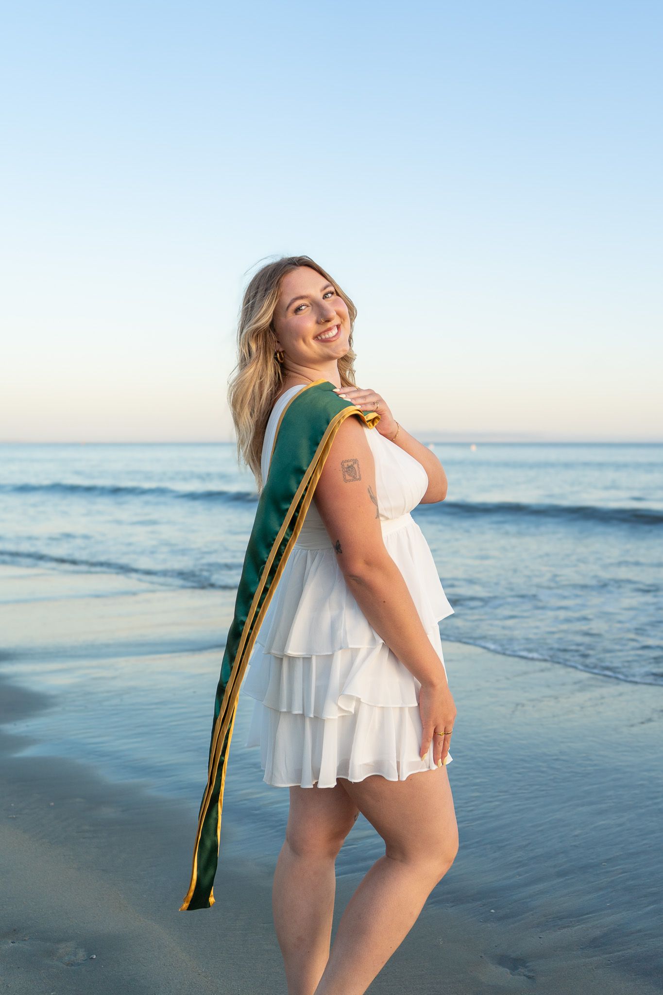 Smiling blonde woman in white dress wearing green graduation sash poses on beach at sunset.