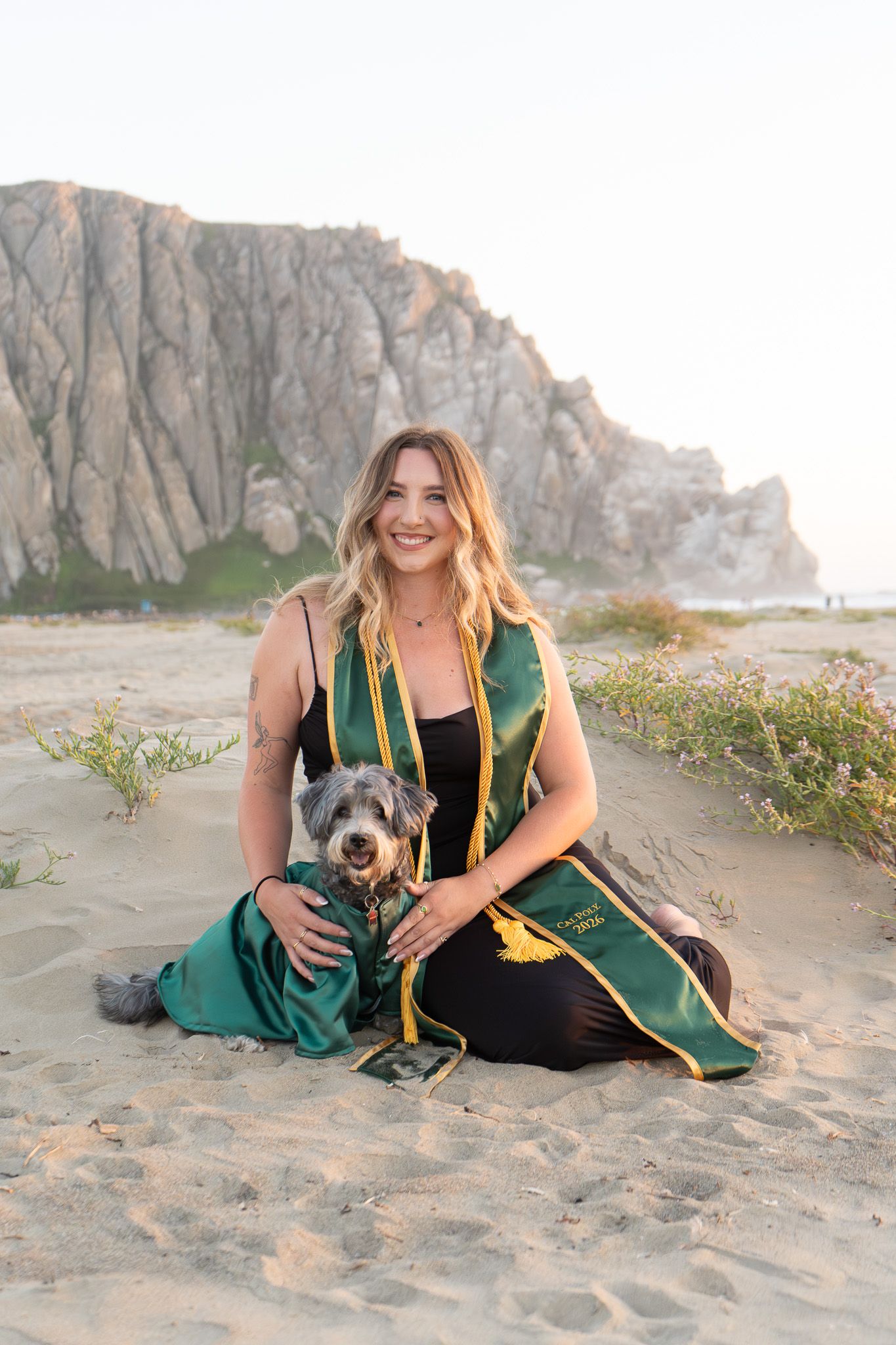 Woman in green graduation stole sits with small dog on sandy beach near rocky cliff.