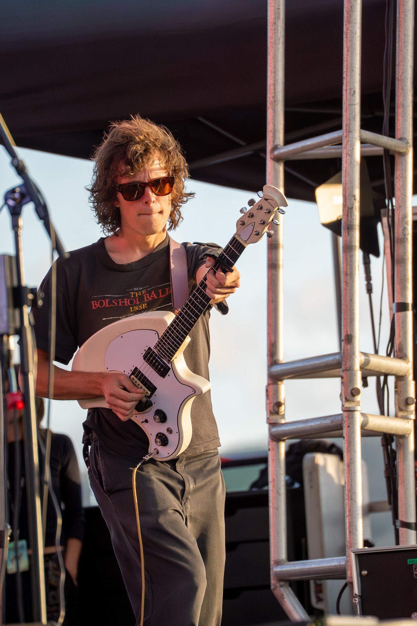 Curly-haired guitarist in sunglasses plays white electric guitar on outdoor stage during live concert performance.