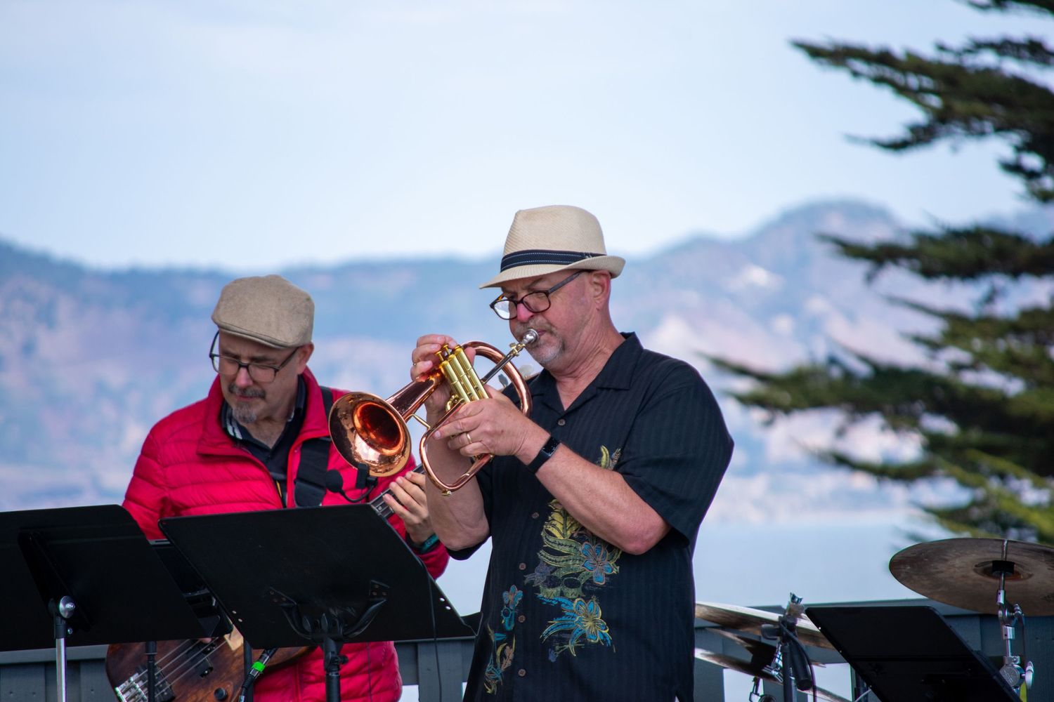 Musicians playing brass instruments perform at outdoor concert with coastal mountains in background.