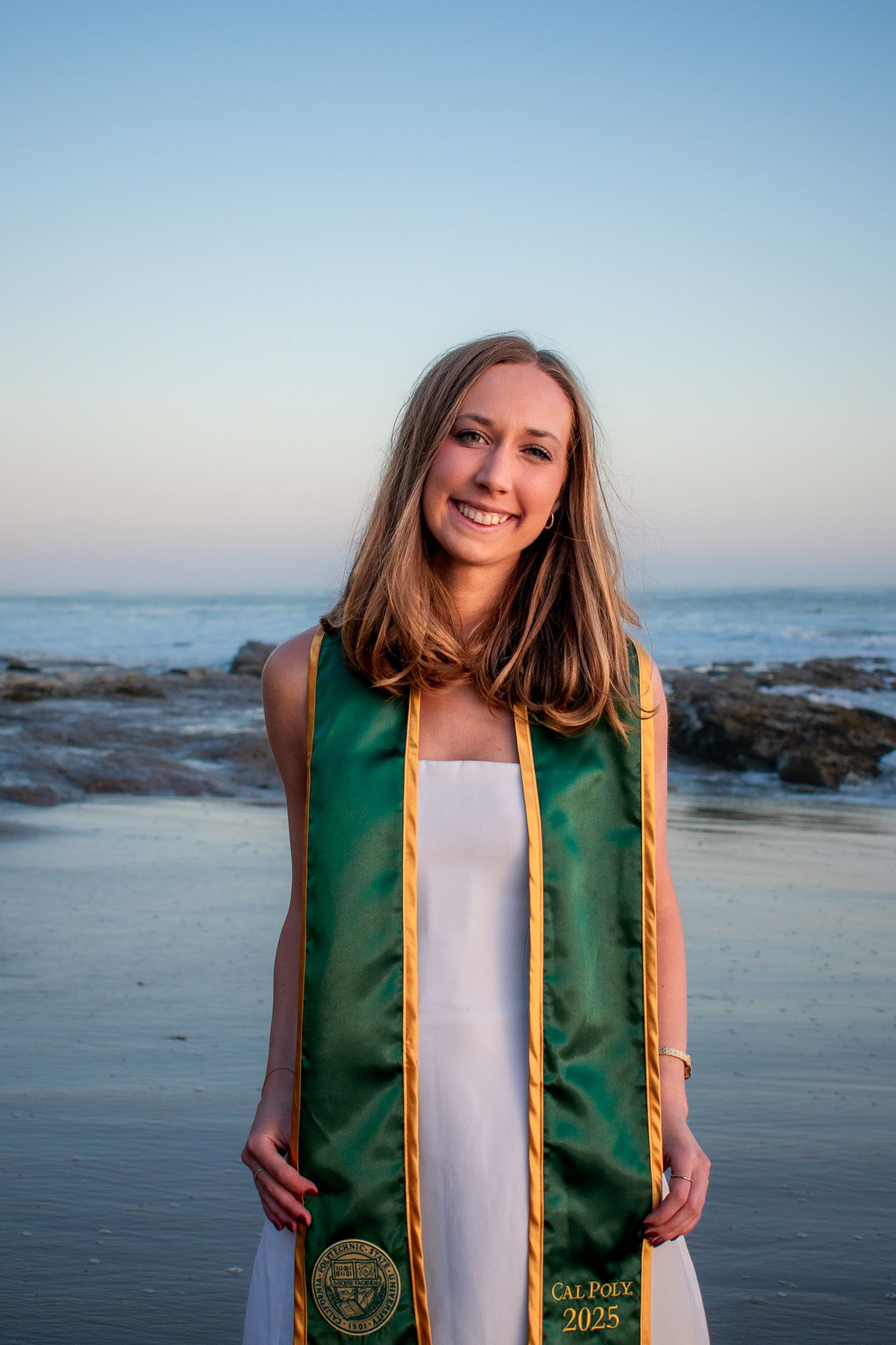 Graduate in green and gold stole poses for photos at sunset on a beach.