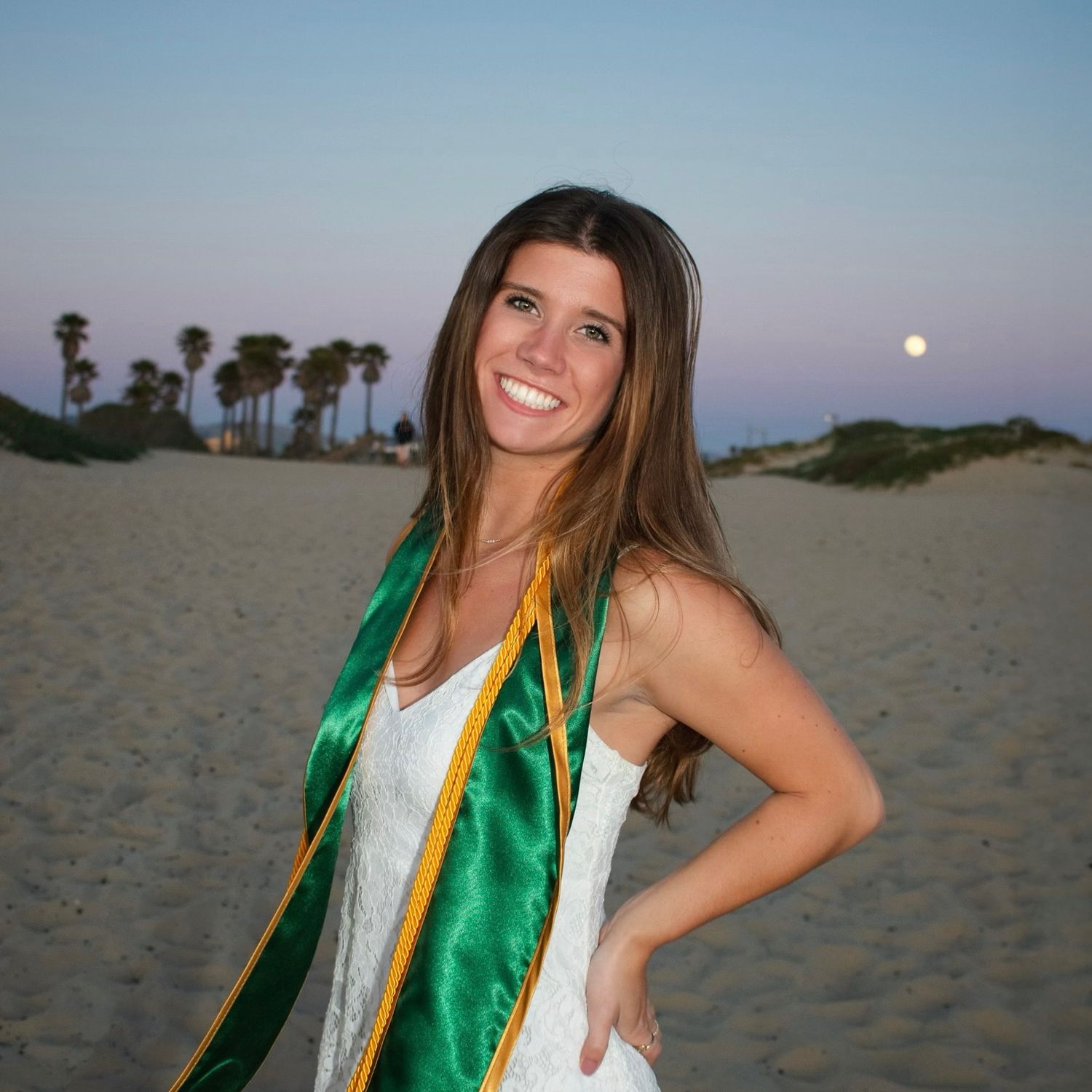 A graduation celebration photoshoot on sandy dunes at sunset with green and gold graduation stoles.