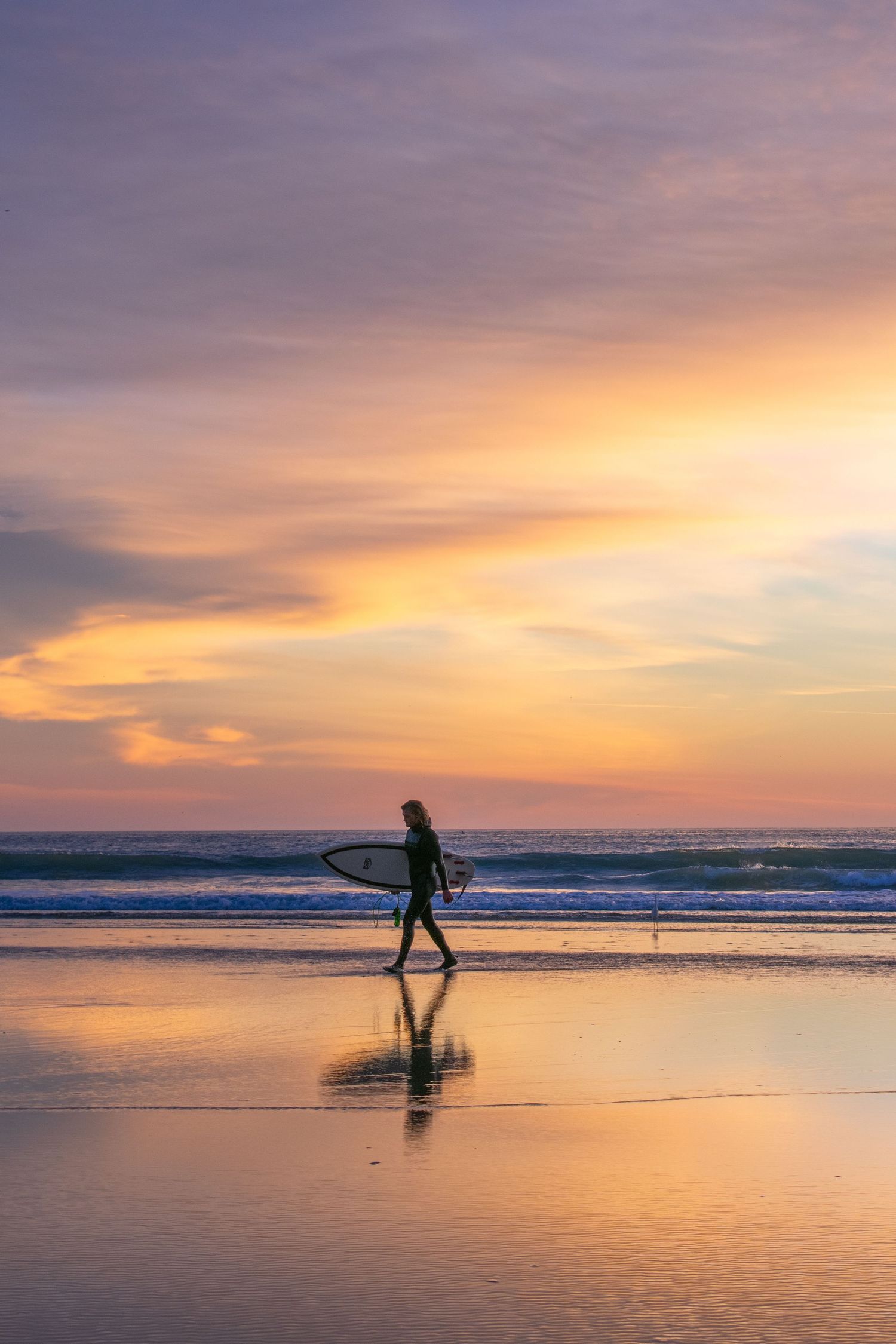 Silhouette of a surfer walking along a beach at sunset with vibrant orange and pink sky.