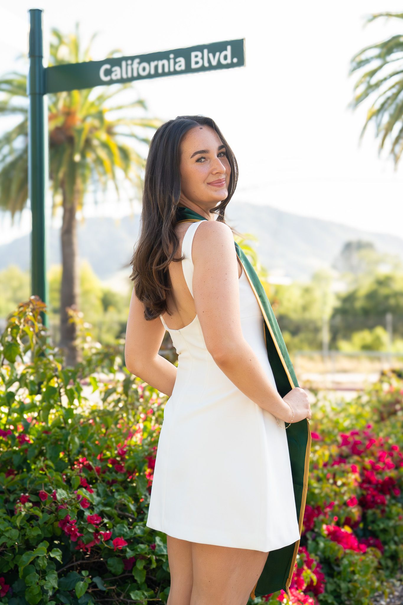 Young woman in white dress holding green graduation sash near California Blvd sign, surrounded by colorful flowers.