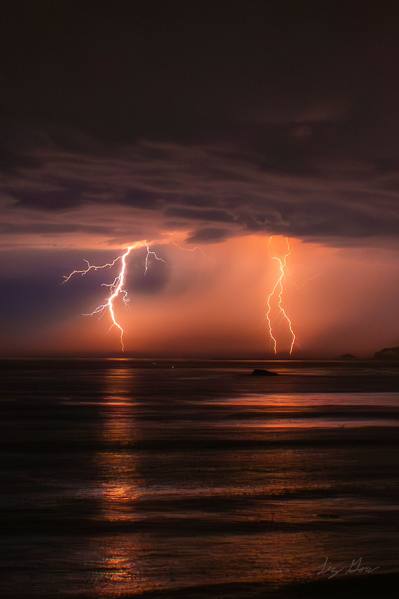 Lightning bolts strike over a dark ocean at sunset creating dramatic reflections on the water's surface.