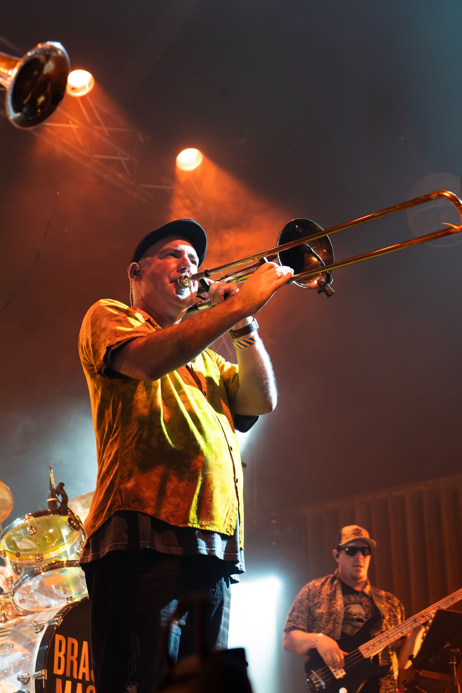 Musician plays trombone on stage in yellow shirt with dramatic orange stage lighting and band members in background.