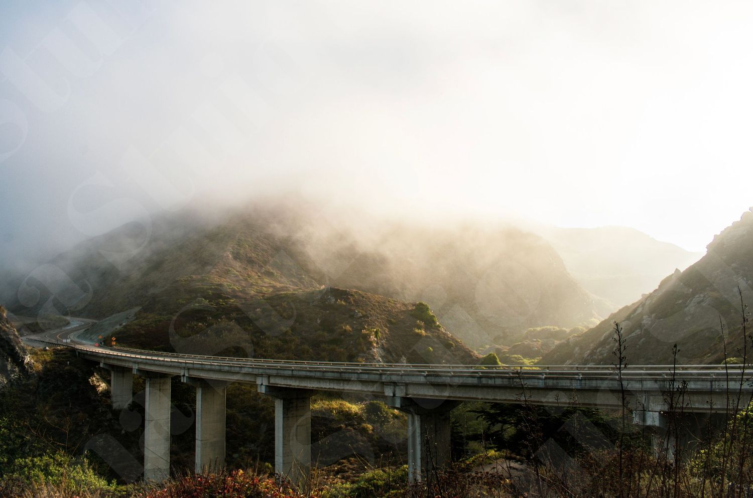 Elevated highway bridge spans across foggy mountain valley in dramatic lighting.