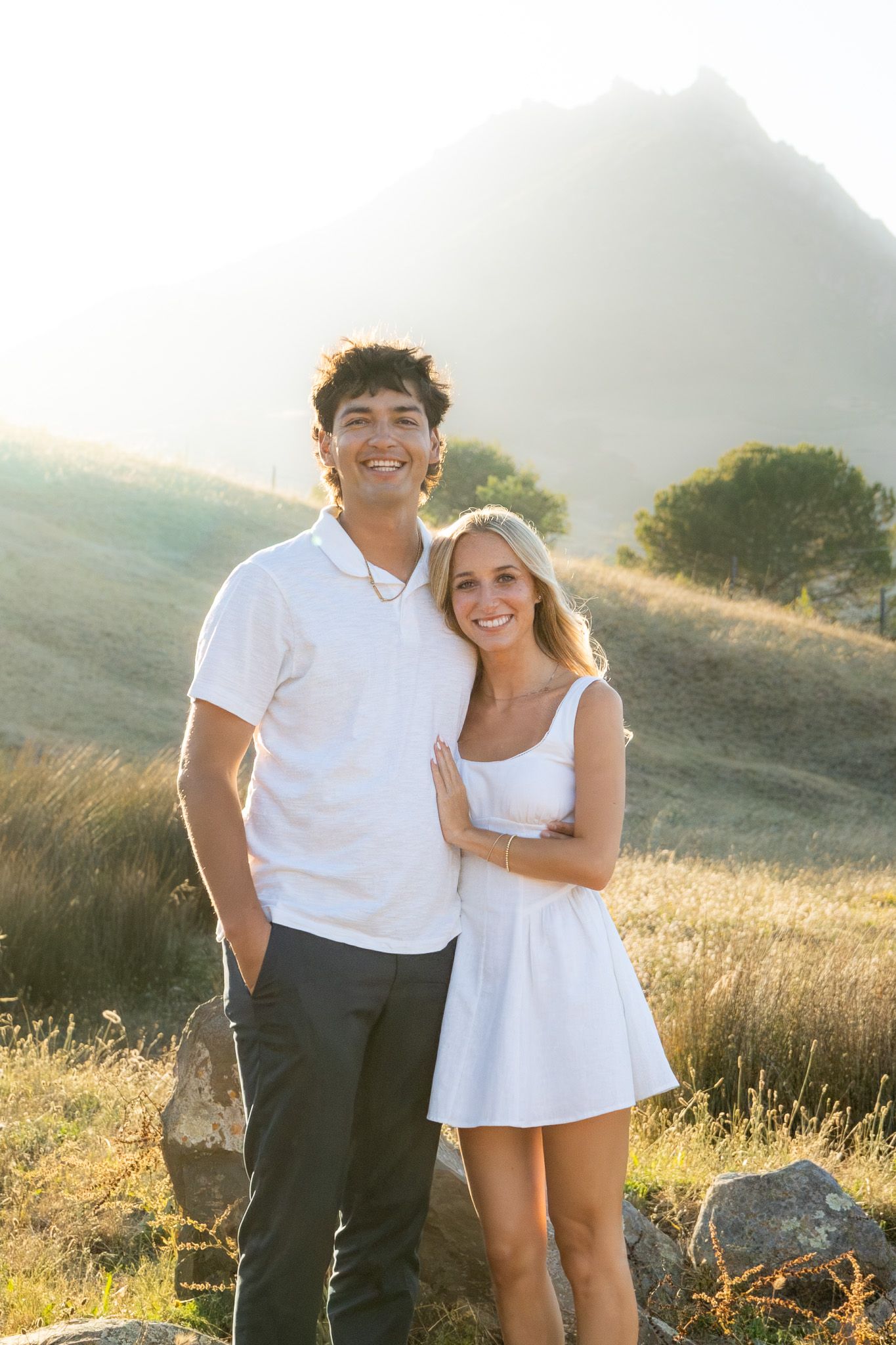 A smiling couple in white outfits poses together in a sunny golden field with a mountain backdrop.