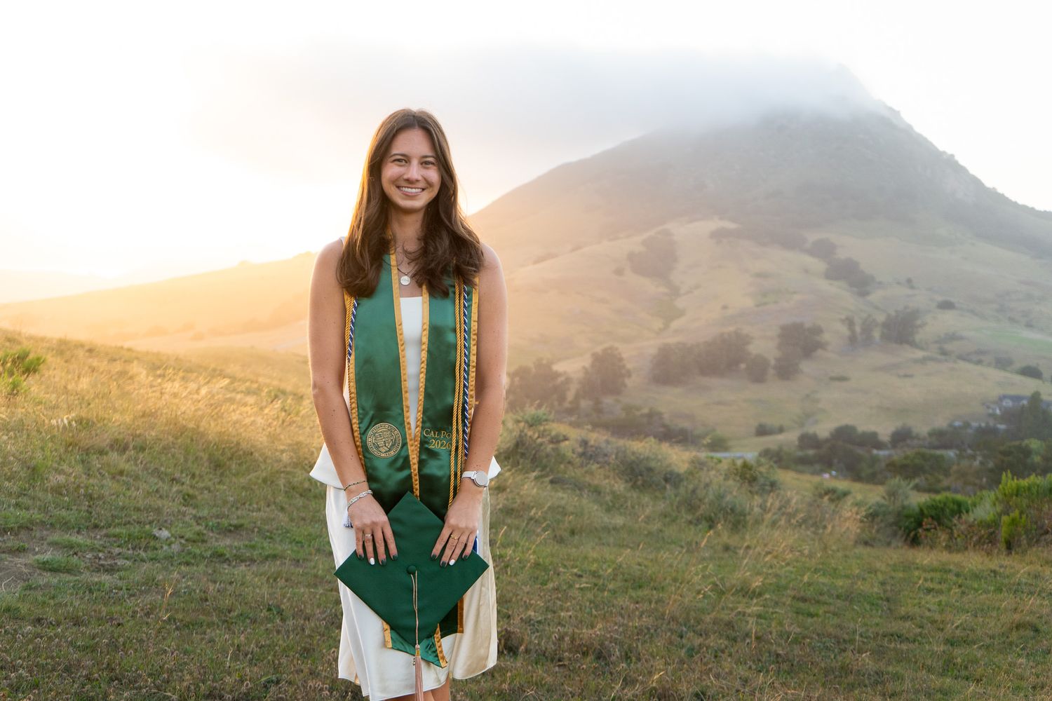 Smiling graduate in green stole and cap poses on hillside at golden hour with scenic mountain backdrop.