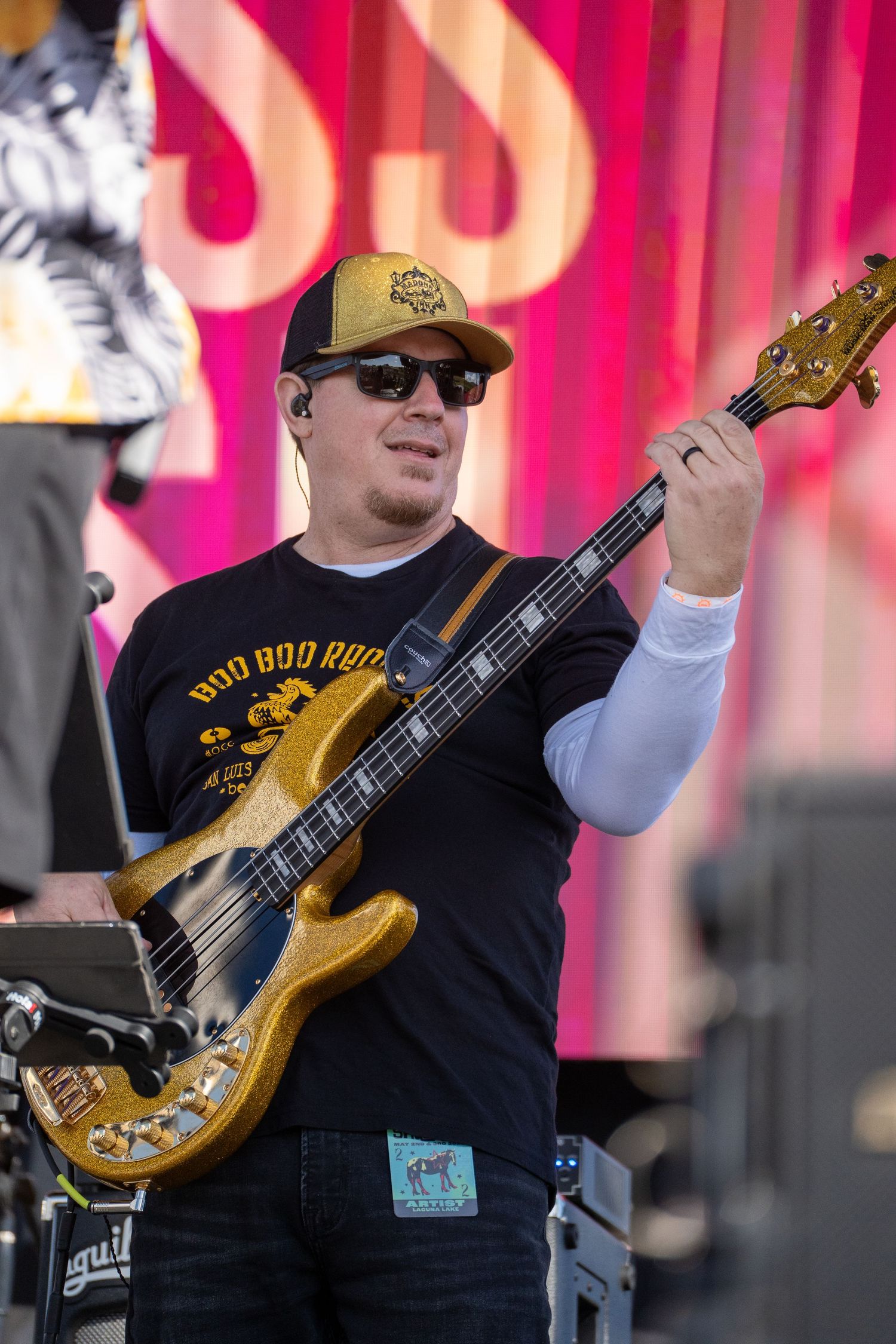 Musician playing a gold bass guitar on stage, wearing sunglasses and a black t-shirt at an outdoor concert.