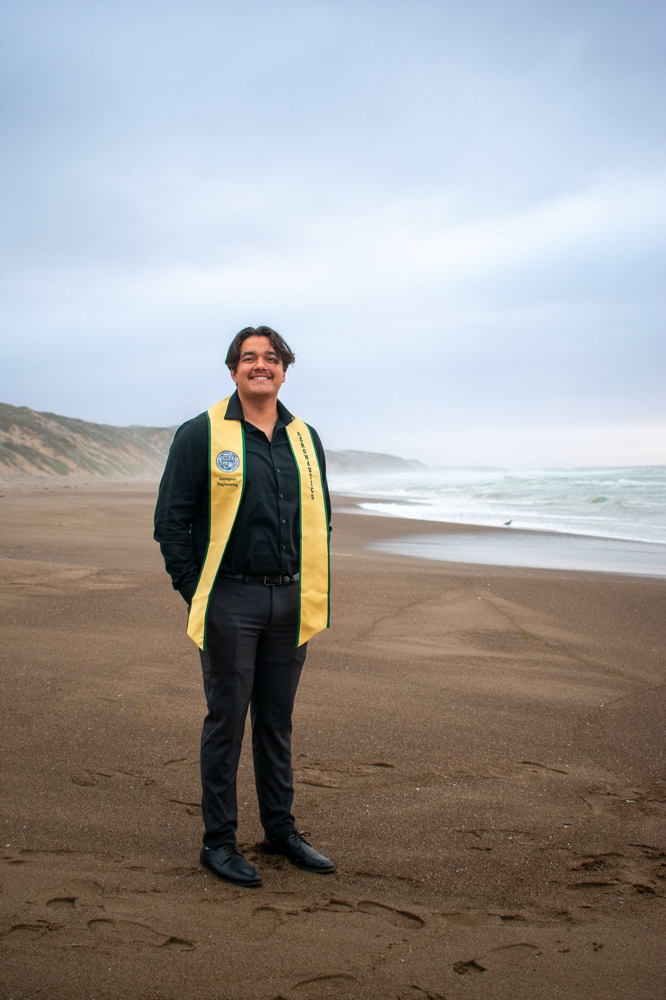 Graduate in black gown and yellow stole stands on foggy beach with mountains in background.