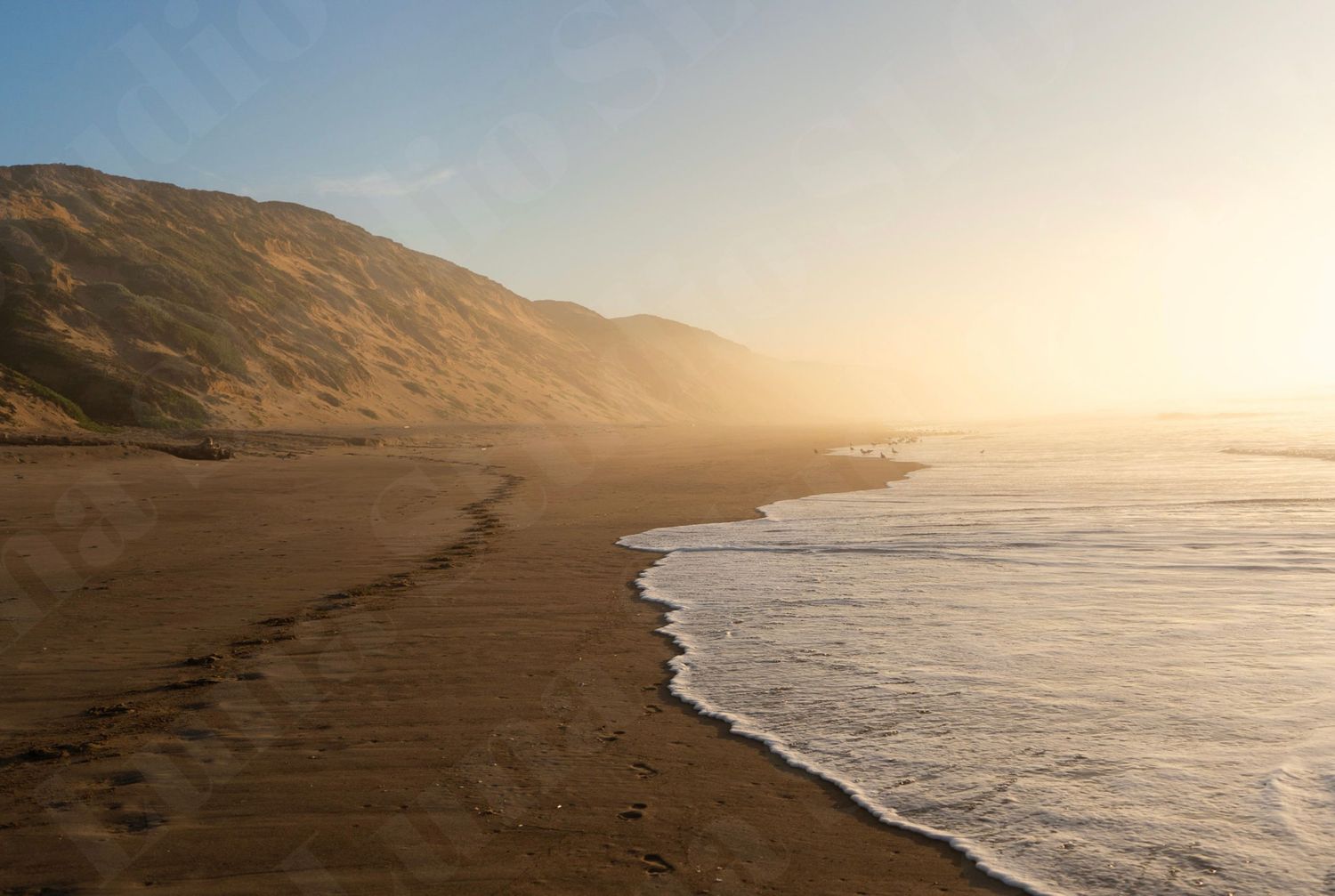 Misty golden sunset over a serene sandy beach with gentle waves and rolling hills along the California coastline.
