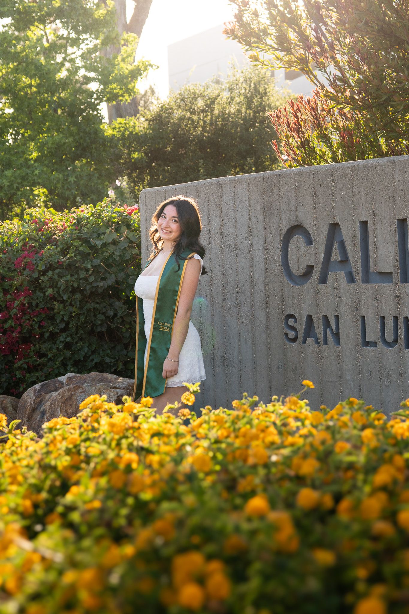 A Cal Poly SLO graduate poses in a white dress and green sash beside the campus sign, surrounded by yellow flowers.