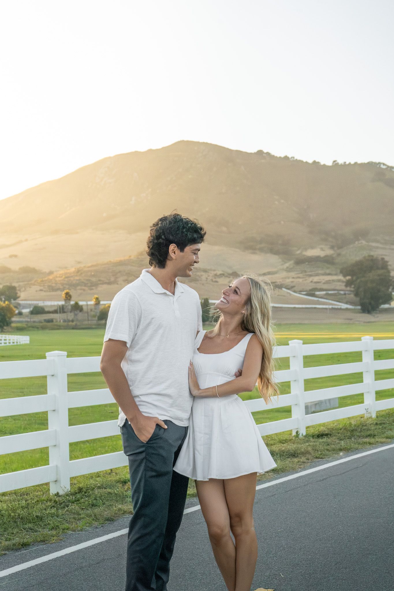 A smiling couple in white outfits stands by a white fence with a sunlit hill and green fields in the background.