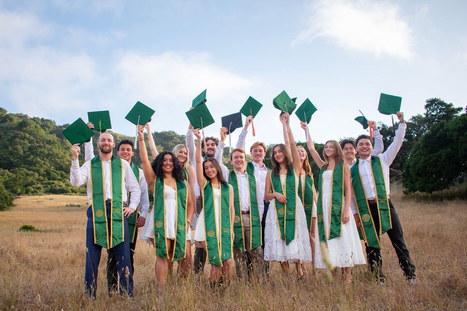 Graduates in white dresses and green stoles celebrate together in an outdoor field during golden hour.