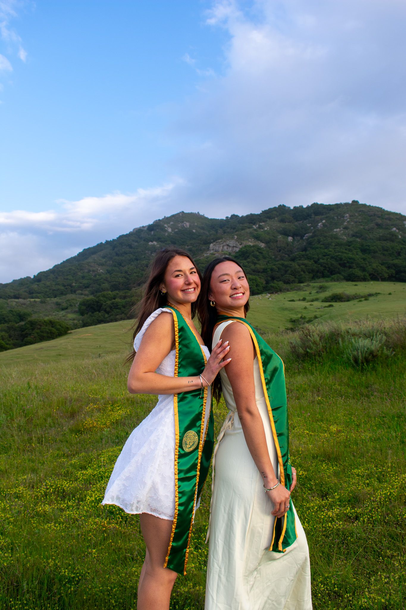 Two graduates in green stoles celebrate in a sunny meadow with hills behind them.